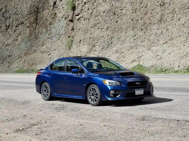 A blue Subaru WRX sedan is parked on a gravel shoulder next to a paved road with a large rocky cliff in the background. The…