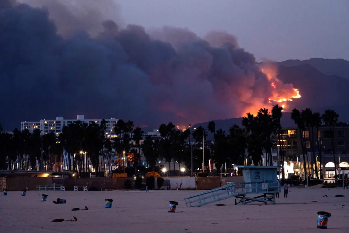 a dramatic photograph shows a massive wildfire burning on distant mountains at dusk, with thick smoke billowing into the sky…