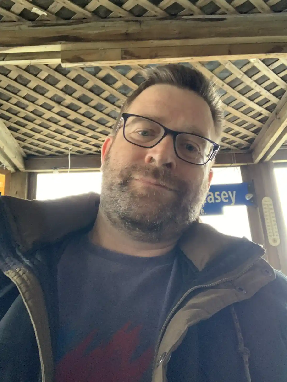 A man with glasses and a beard takes a selfie under a wooden lattice roof, wearing a jacket over a t-shirt. Behind him, a…