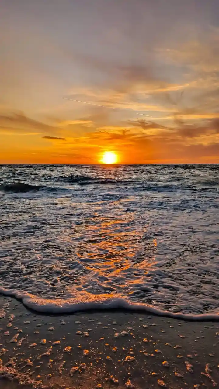 A serene beach sunset photo with the sun low on the horizon, casting orange and yellow light across the sky and reflecting…