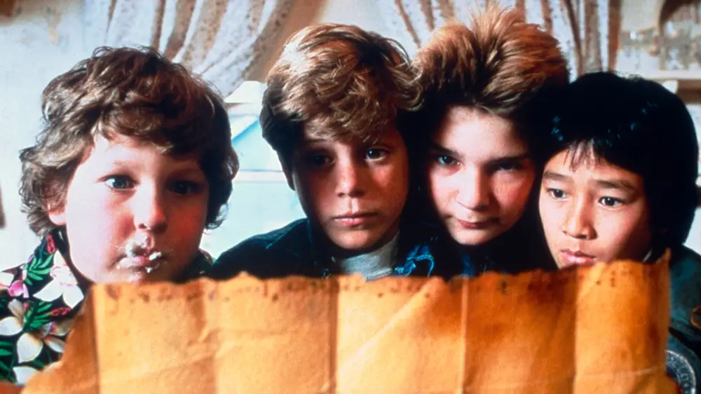 four young boys from the 1980s movie 'The Goonies' peer over a torn brown paper bag, their faces showing intense…