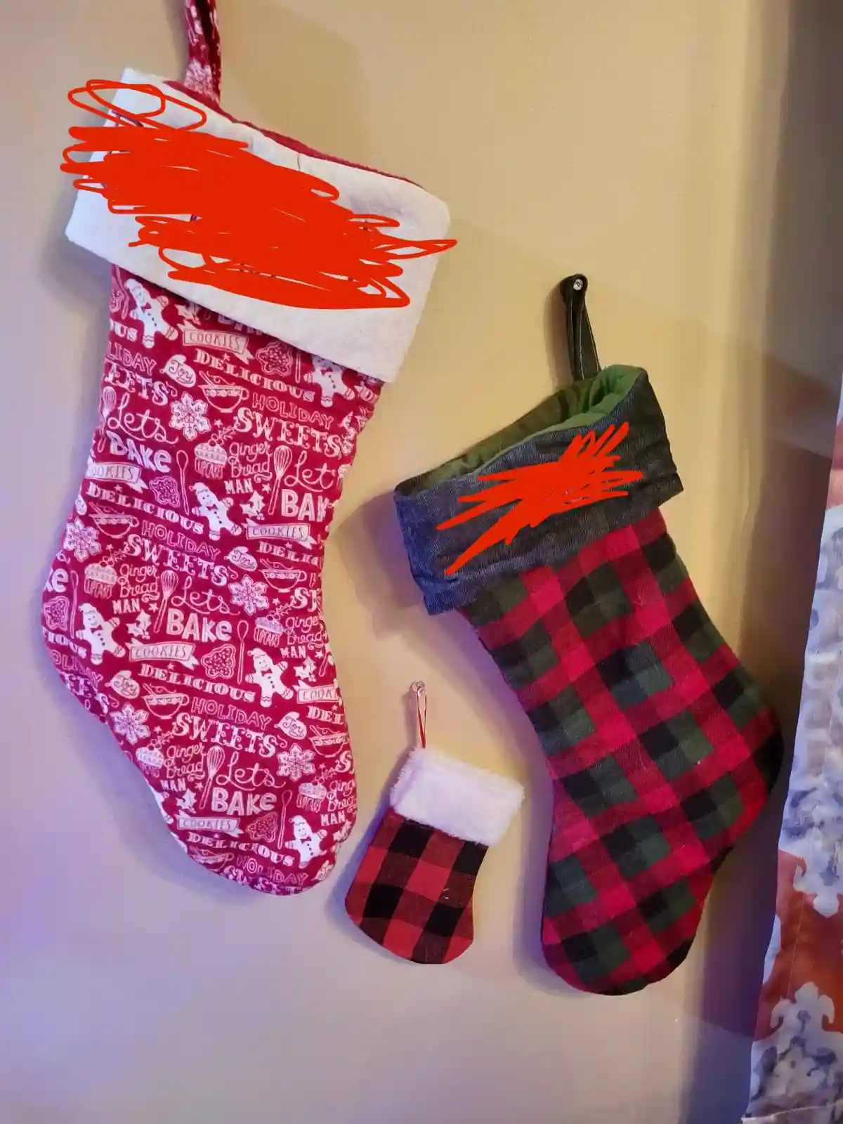 Three Christmas stockings hang on a wall, two large and one small, all featuring festive patterns. The larger red stocking…