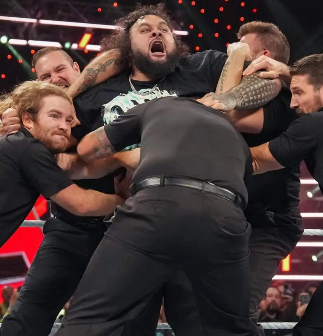 A large, bearded wrestler with tattoos is being held down by multiple men in black shirts inside a wrestling ring, his mouth…