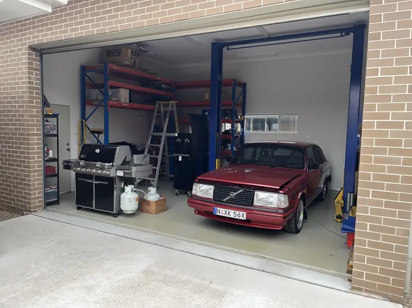 A red Volvo sedan is parked inside an open garage next to a large stainless steel barbecue grill with propane tanks. The…