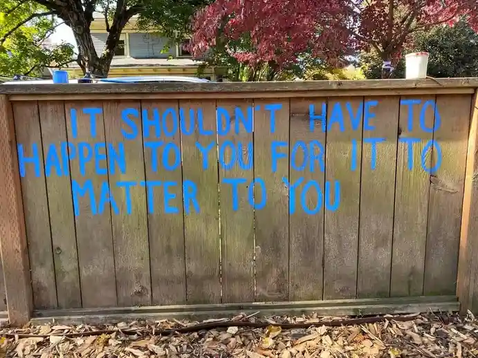 A wooden fence in a residential yard has blue spray-painted text that reads 'IT SHOULDN'T HAVE TO HAPPEN TO YOU FOR IT TO…