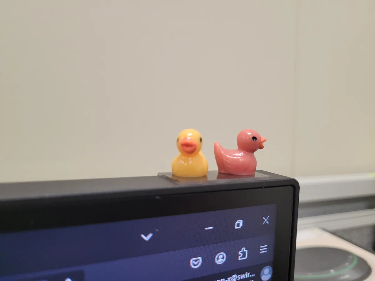 Two small rubber ducks, one yellow and one pink, sit side-by-side on top of a black electronic device with a visible…