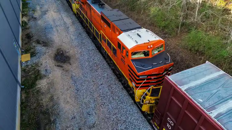 An aerial view shows an orange and yellow Genesee & Wyoming locomotive, number 4055, stopped on a gravel track next to a red…