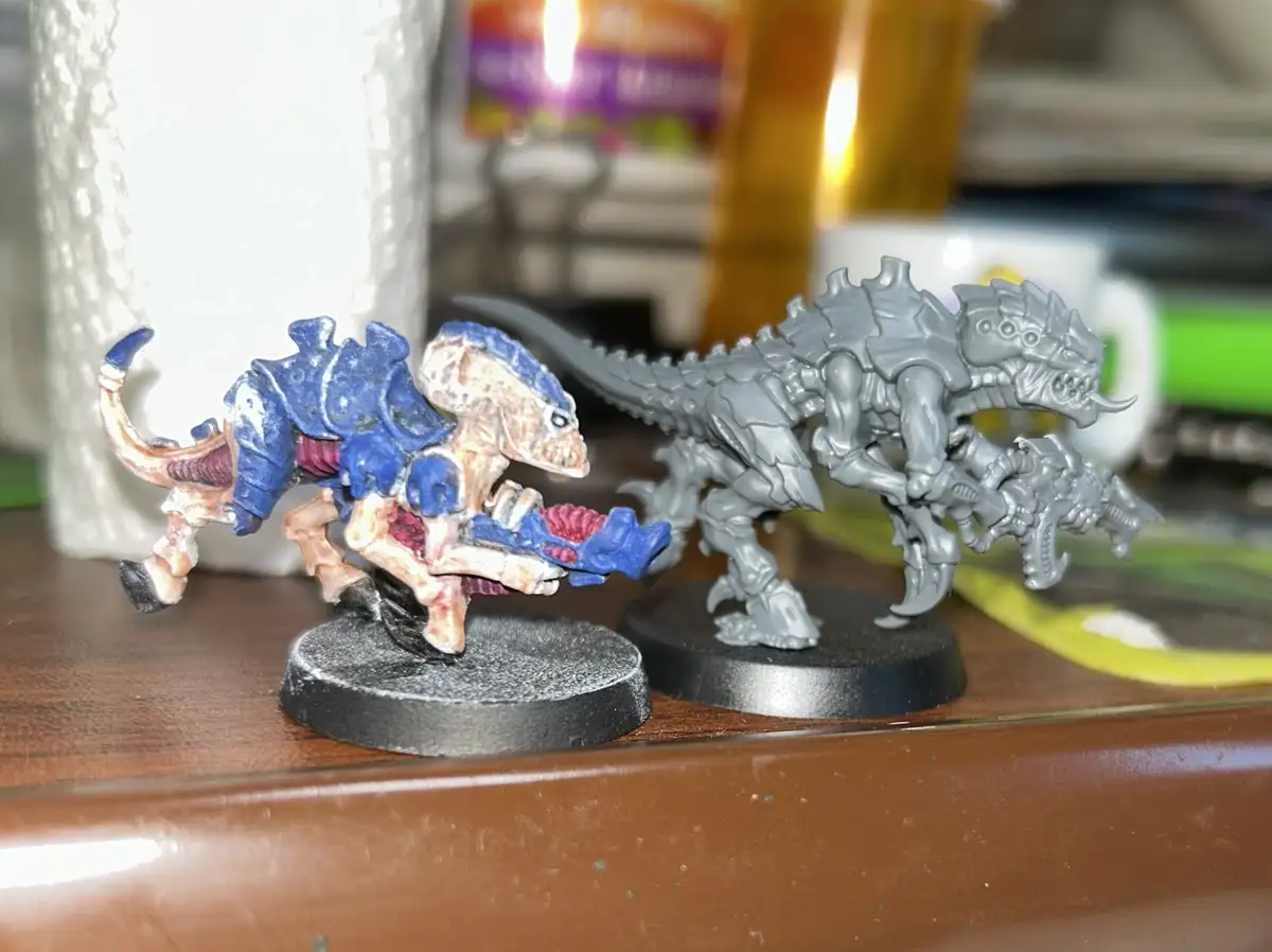 Two miniature fantasy creatures sit side-by-side on a wooden surface; the one on the left is painted with blue, pink, and…