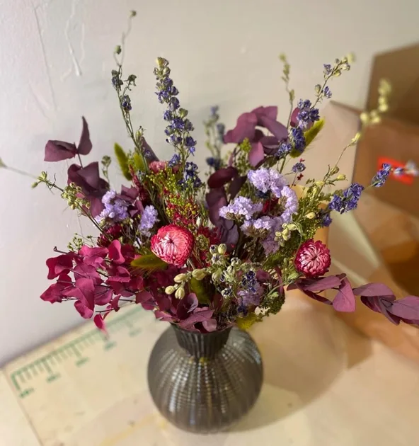 A vibrant bouquet of dried flowers in purples, pinks, and blues sits in a textured gray vase on a wooden surface. A green…