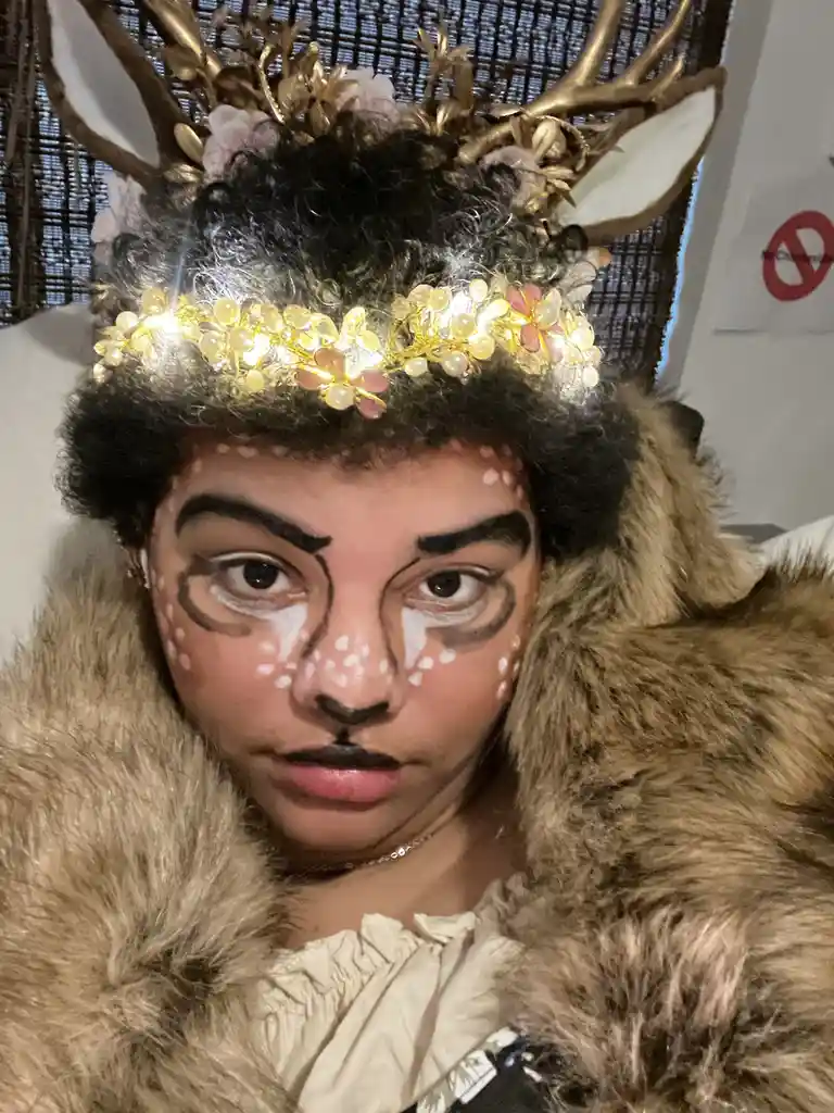 A person with dark curly hair wears a deer costume featuring golden antlers with white tips, a glowing floral headband, and…