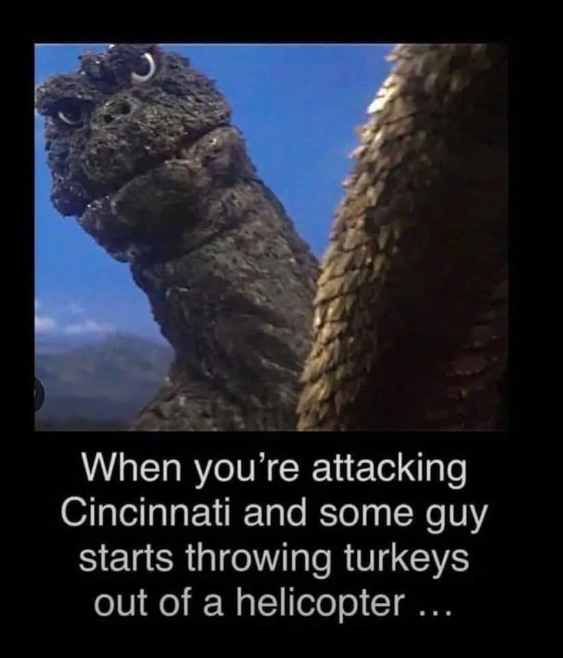 A close-up shot of the kaiju Godzilla, showing his textured, scaly head and neck against a blue sky, with his tail visible…