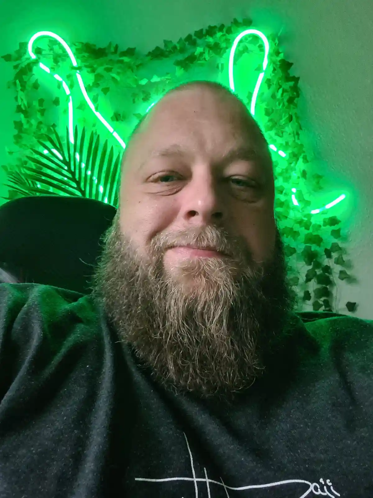 A man with a full beard and shaved head smiles at the camera while sitting in a chair. Behind him, a green neon light shaped…