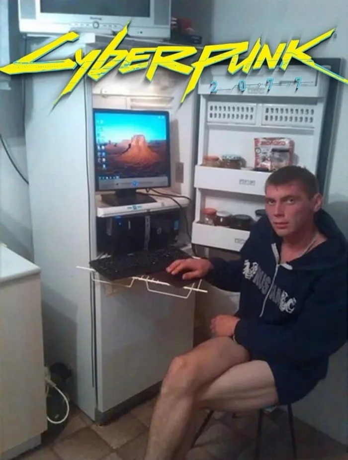 A man sits on a chair in a cramped room, using a computer setup that's literally built into a refrigerator. The monitor is…