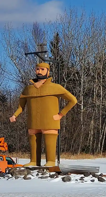 A large, stylized statue of a man in a yellow outfit stands on a snowy landscape with bare trees in the background. The…