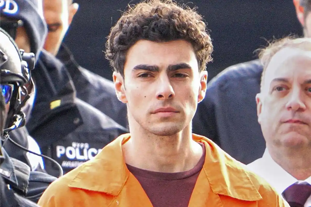 A man with curly dark hair wearing an orange prison jumpsuit stands solemnly, flanked by NYPD officers and another man in a…