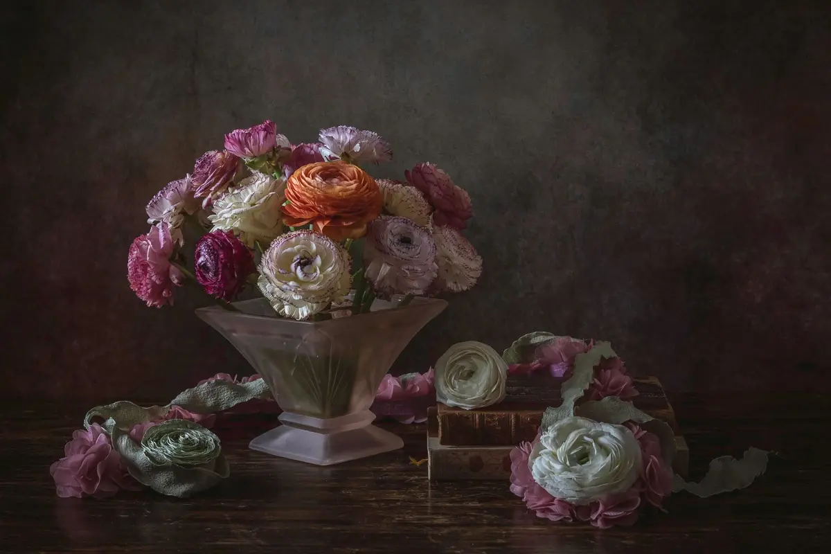 A still life photograph shows a bouquet of ranunculus flowers in a clear glass vase, with additional flowers and petals…