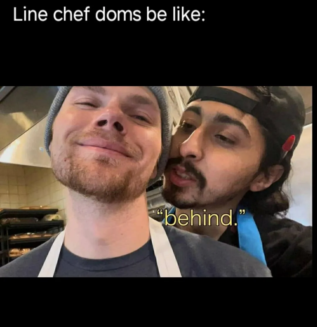 Two men in a kitchen setting, one in the foreground wearing a beanie and apron with a smug, closed-eye smile, while the…