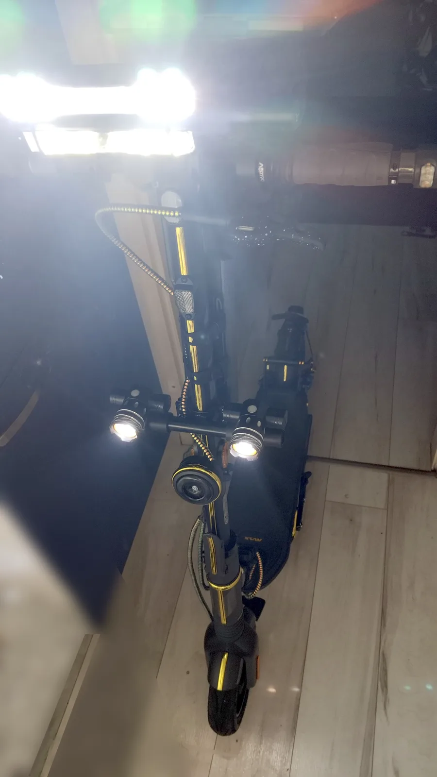 A black and gold electric scooter with multiple bright lights is shown from a high angle indoors on a light wood floor. The…