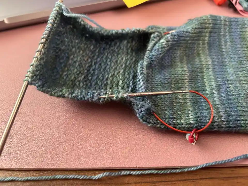 A close-up photo shows a partially knitted sock in muted green and blue yarn, resting on a pink textured surface. Metal…