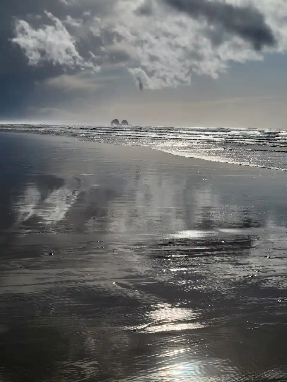 A wide-angle photo of a wet, reflective beach under a cloudy sky, with waves lapping at the shore and distant rock…