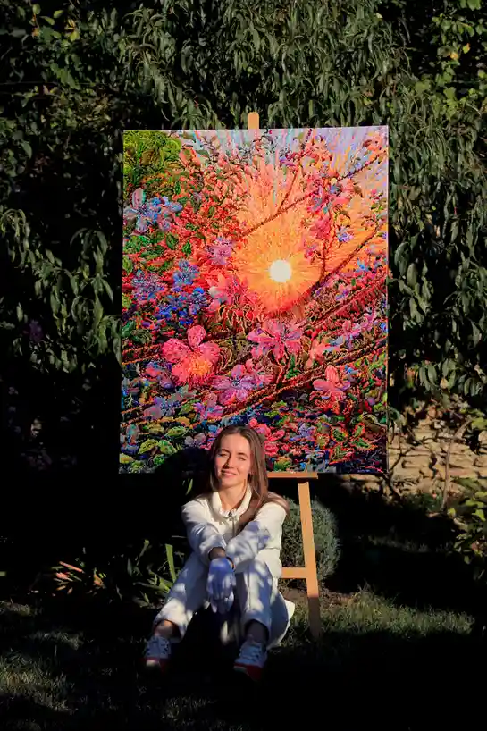 A young woman in a white outfit and gloves sits on the grass in front of a large, vibrantly colorful painting on an easel…