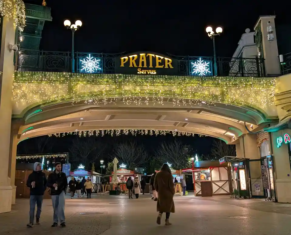 A nighttime photo of the entrance to the Prater amusement park in Vienna, decorated with festive string lights and…