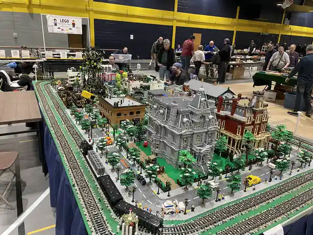 A large, intricate LEGO cityscape built on a table at a convention, featuring detailed buildings, trees, roads, and…
