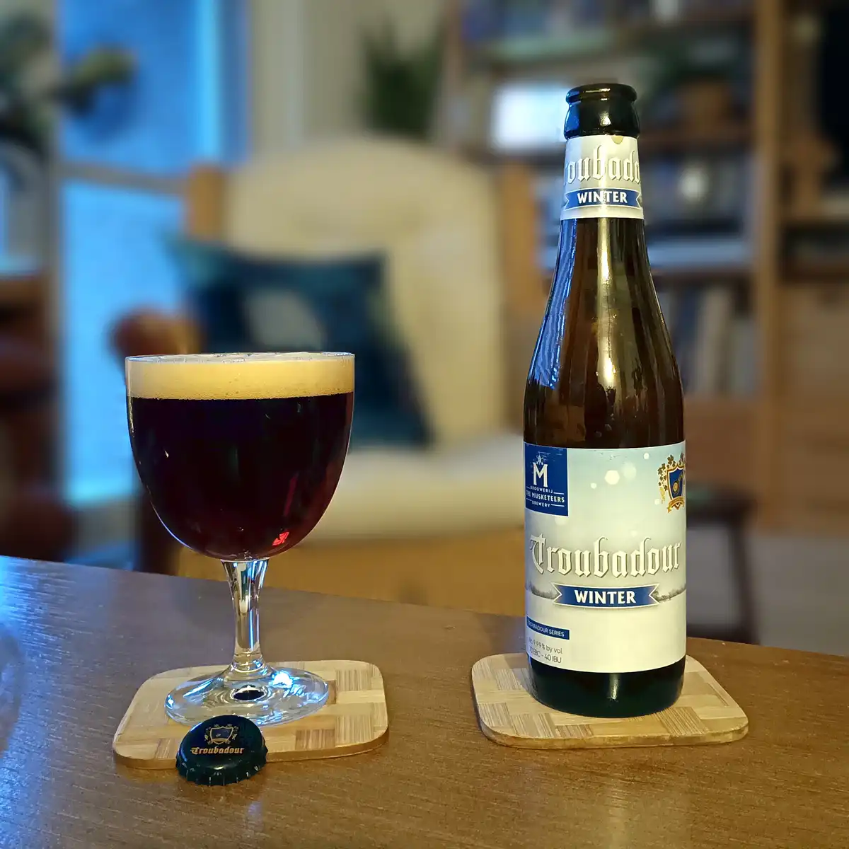 A bottle and glass of Troubadour Winter beer sit on wooden coasters on a table, with the bottle's label and the glass's dark…