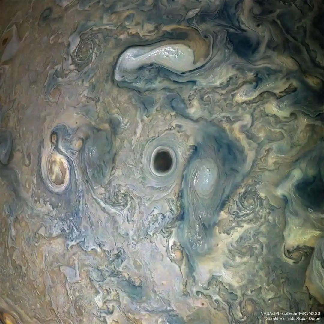 A NASA image of Jupiter's turbulent cloud bands, where swirling patterns of beige, blue, and white gases form a face-like…