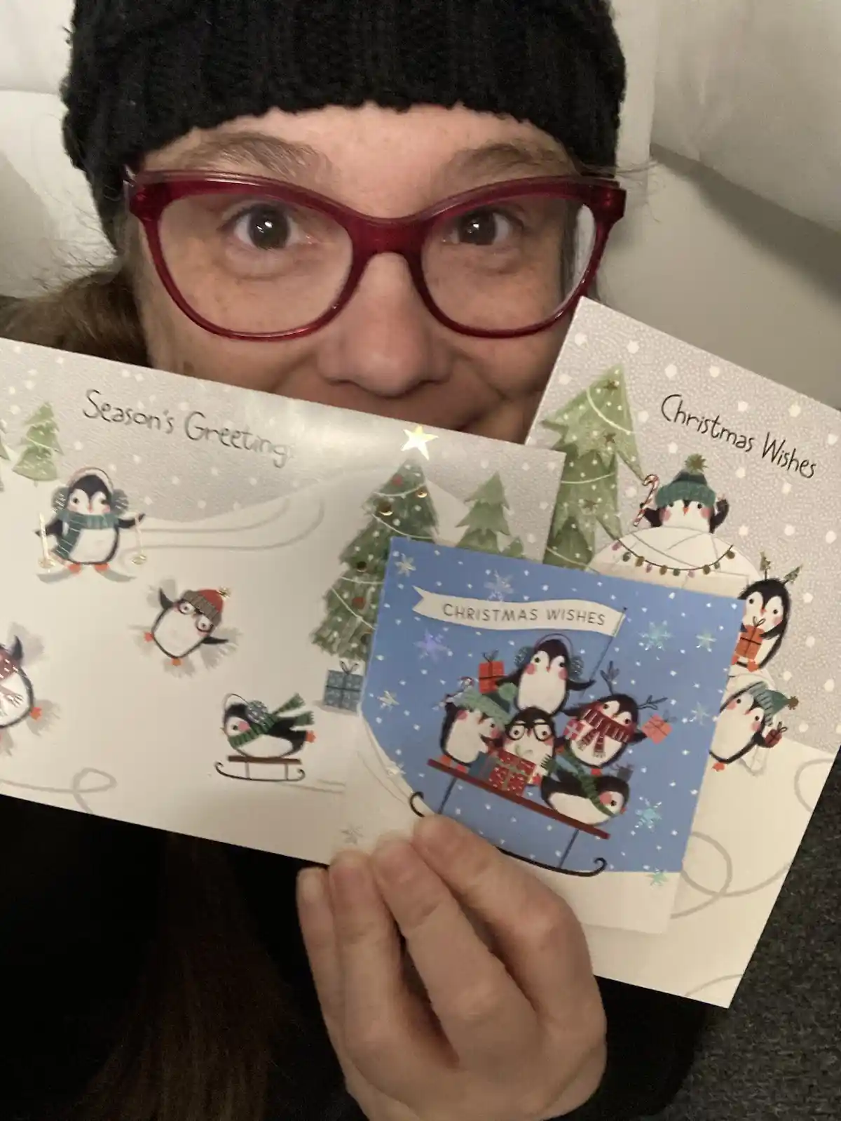 A person wearing red glasses and a black beanie holds up three Christmas cards featuring cartoon penguins in winter scenes…