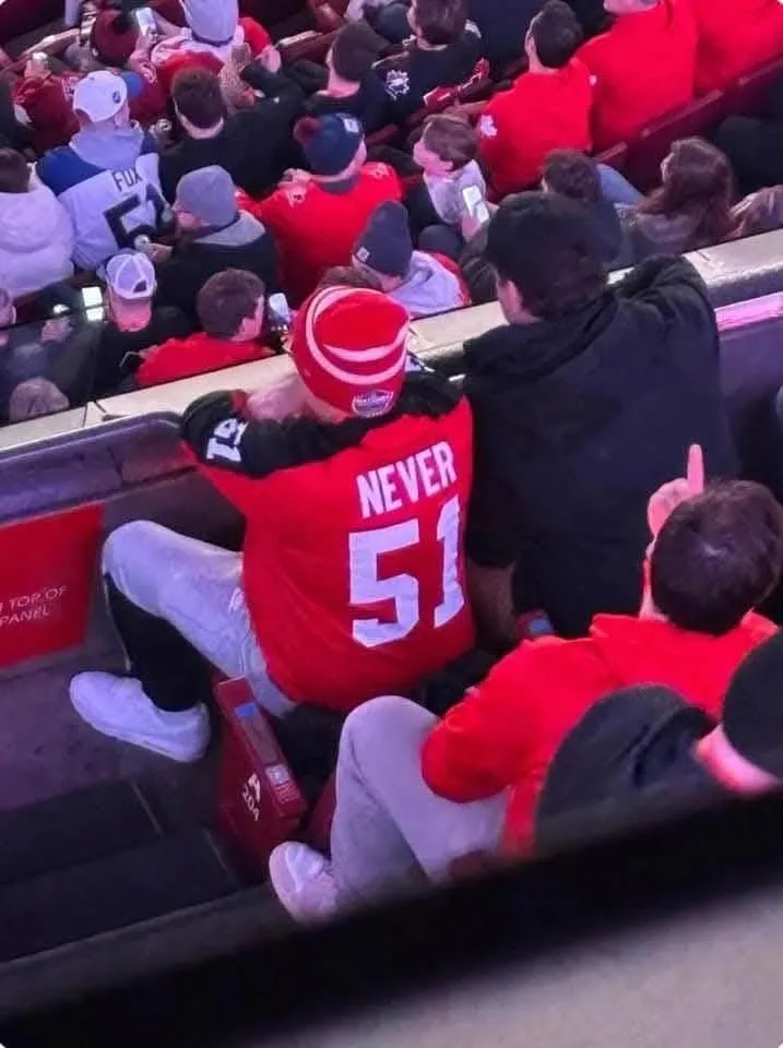 A person wearing a red sports jersey with 'NEVER 51' printed on the back is sitting in a stadium crowd, leaning over the…