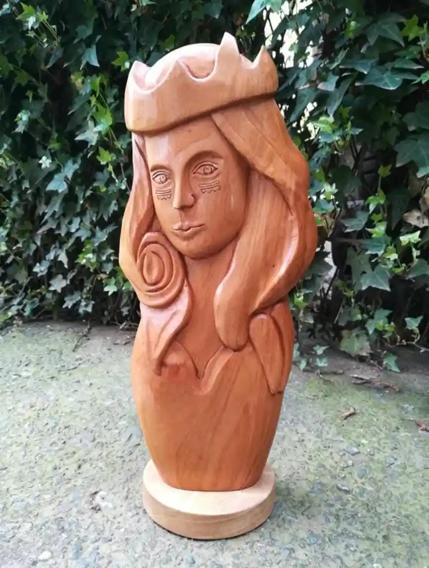 A wooden sculpture of a female figure with long hair, a crown, and a rose carved into her shoulder, standing on a pedestal…