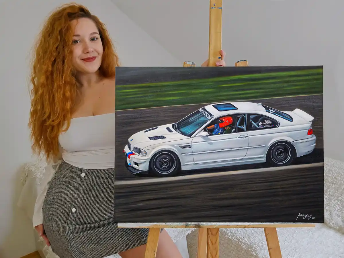 A woman with long curly red hair stands beside an easel displaying a detailed painting of a white BMW M3 race car speeding…