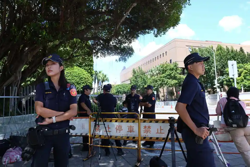 一群穿著深藍色制服的警察站在戶外，背景是綠樹和現代建築，前景有黃黑相間的路障，上面寫著「禁止停車」。警察們表情嚴肅，有的雙臂交叉，有的手持裝備，似乎在執行勤務。路障旁邊有攝影機架設在三腳架上，暗示可能有媒體在現場。整體畫面呈現出一種秩序井然但略帶緊張…