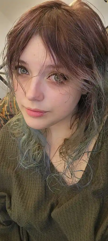 A close-up selfie of a young woman with messy, multi-toned hair (brown, pink, and greenish-blue) and light green eyes…