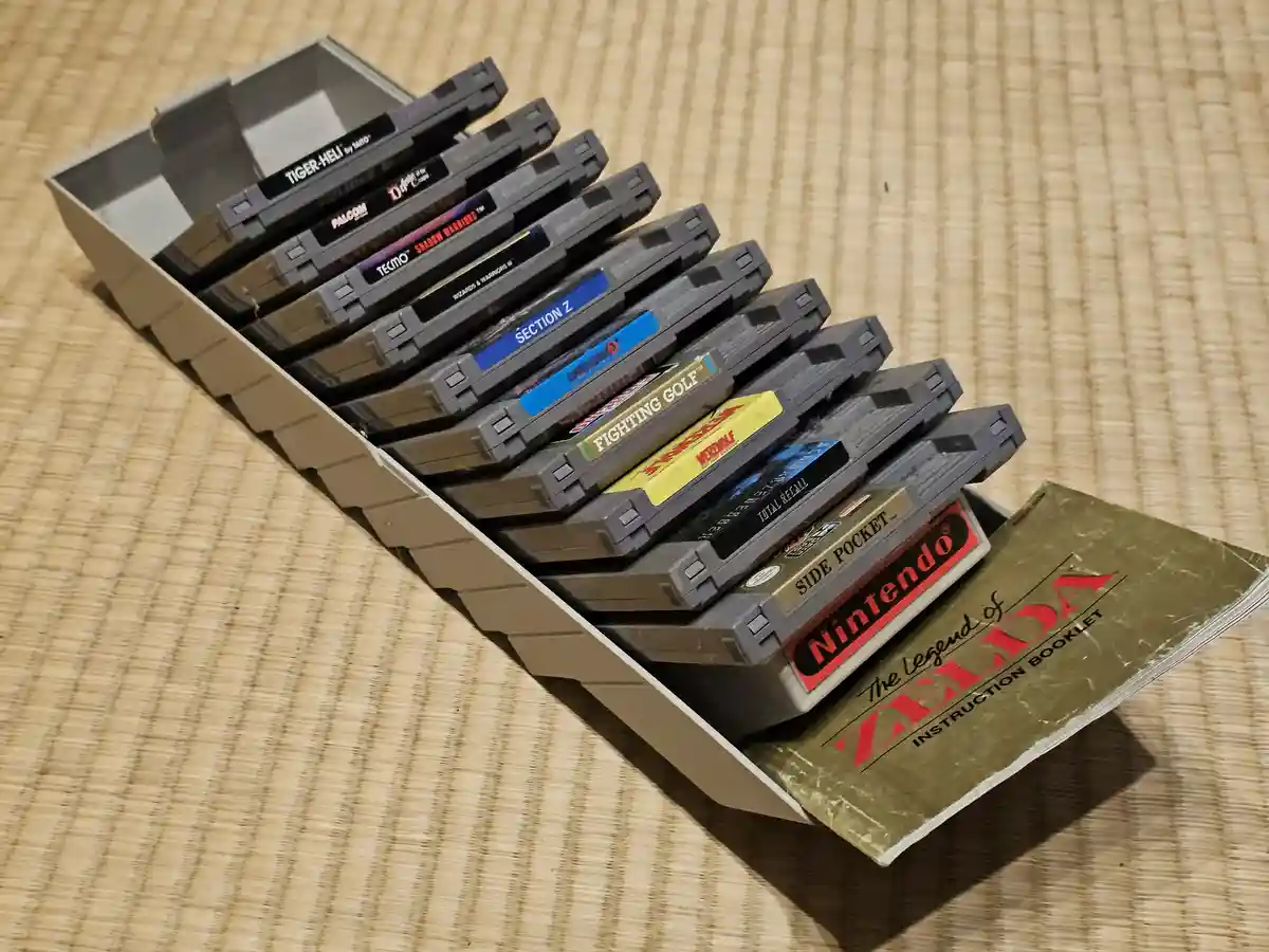 A beige NES game cartridge tray holds a stack of gray NES cartridges, including titles like 'Tiger-Heli', 'Shadow Warriors'…
