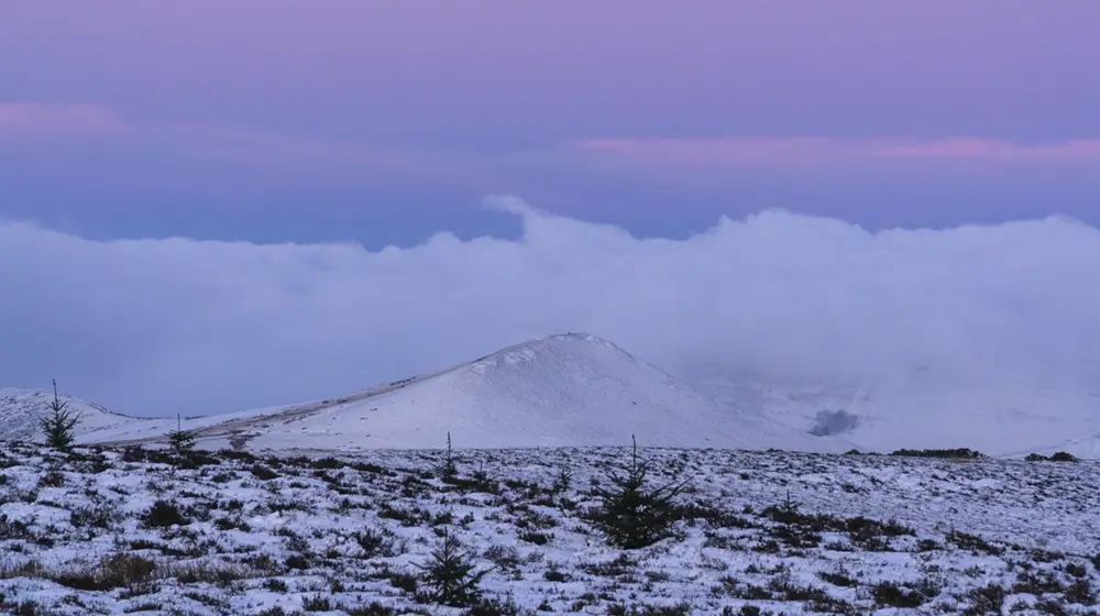 A snowy mountain landscape under a purple and blue twilight sky, with clouds partially obscuring the peak. The foreground…