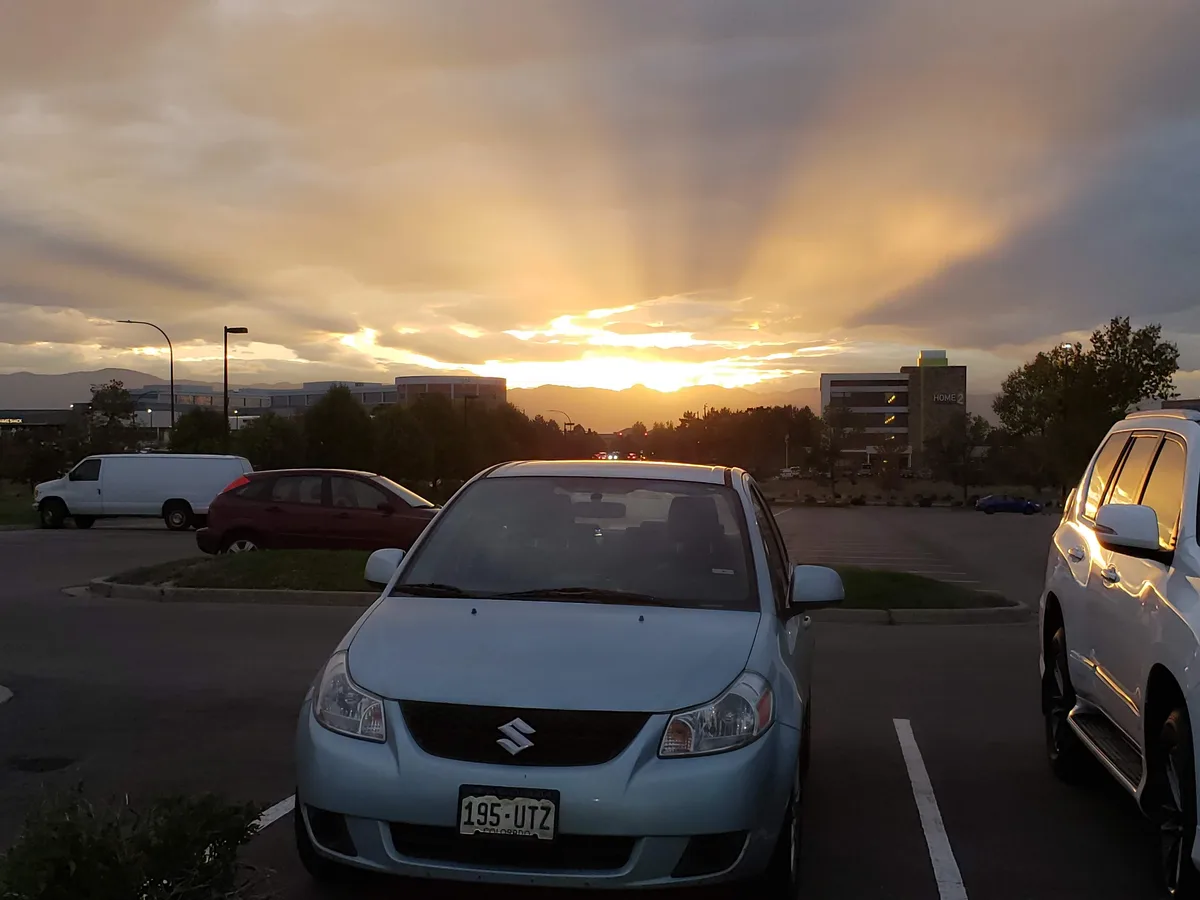 A light blue Suzuki car is parked in a lot during a dramatic sunset with golden rays breaking through clouds. Other cars are…