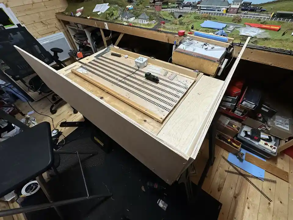 An overhead view of a cluttered hobby room with a partially built model train layout on a wooden workbench. The layout…