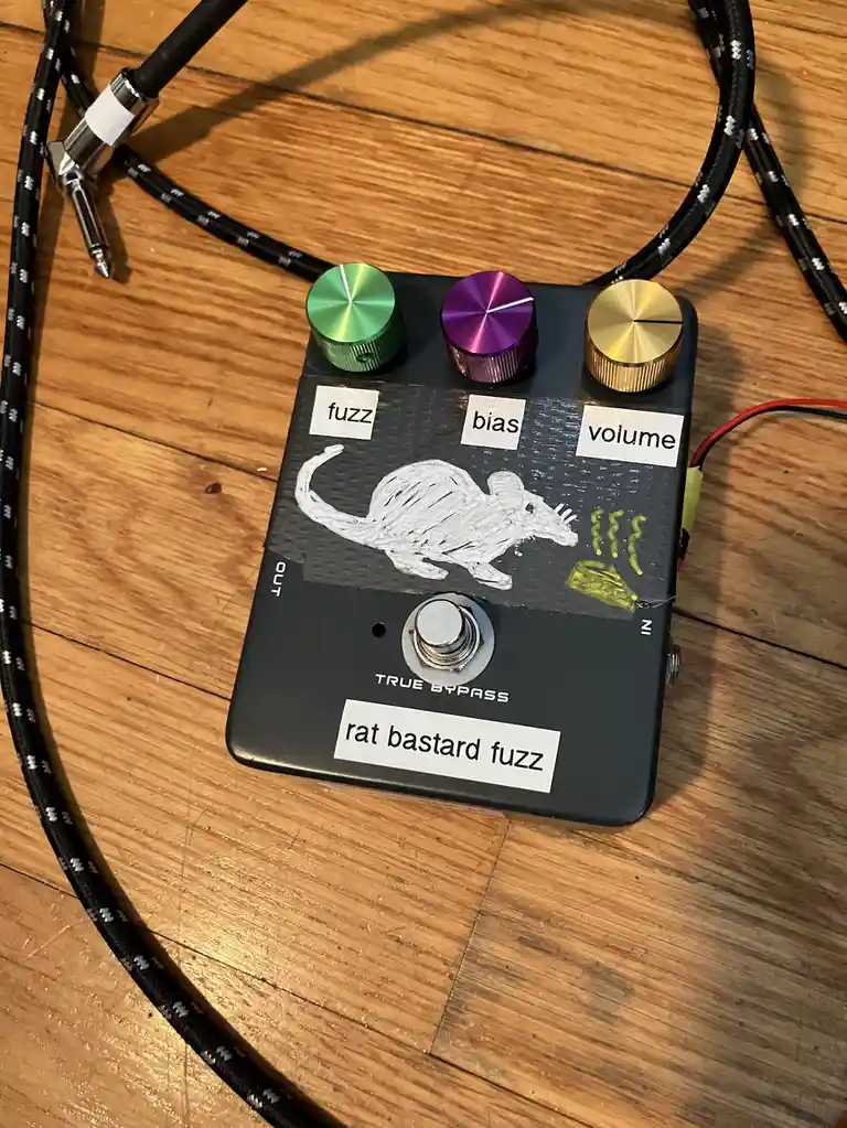 A black guitar effects pedal labeled 'rat bastard fuzz' sits on a wooden floor, featuring a hand-drawn white rat graphic and…
