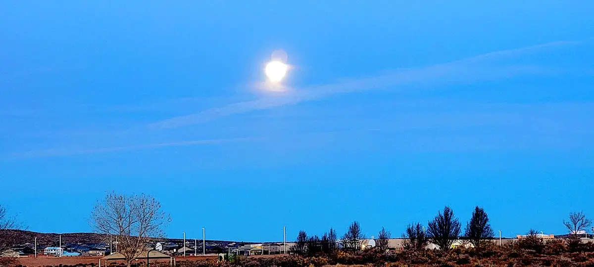 A wide shot of a bright moon glowing in a clear blue sky above a distant suburban landscape with houses, trees, and power…