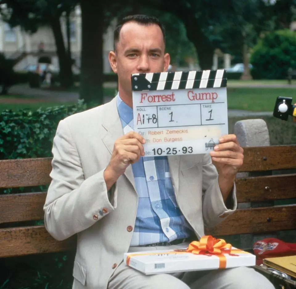 Tom Hanks as Forrest Gump sits on a park bench holding a clapperboard that reads 'Forrest Gump' with scene and take numbers…