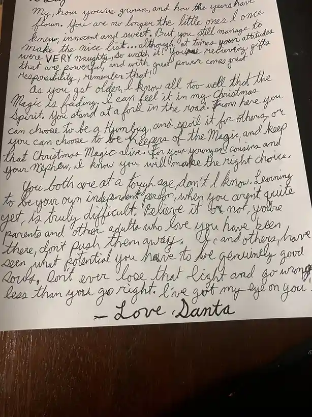 A handwritten letter on white paper, resting on a dark wooden surface, addressed to children by Santa. The cursive script is…