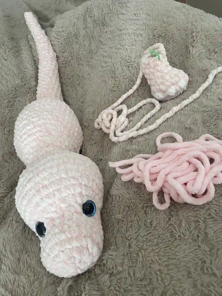 A handmade pink and white crocheted snake plushie with large blue eyes lies on a gray fluffy blanket, next to a pile of…