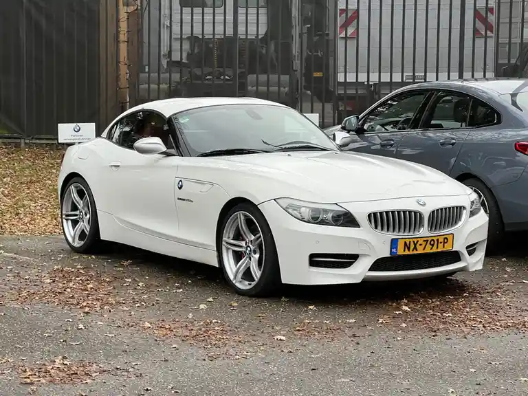 A white BMW Z4 convertible is parked on a leaf-strewn asphalt lot next to a gray sedan, with a sign reading 'Parkeren…