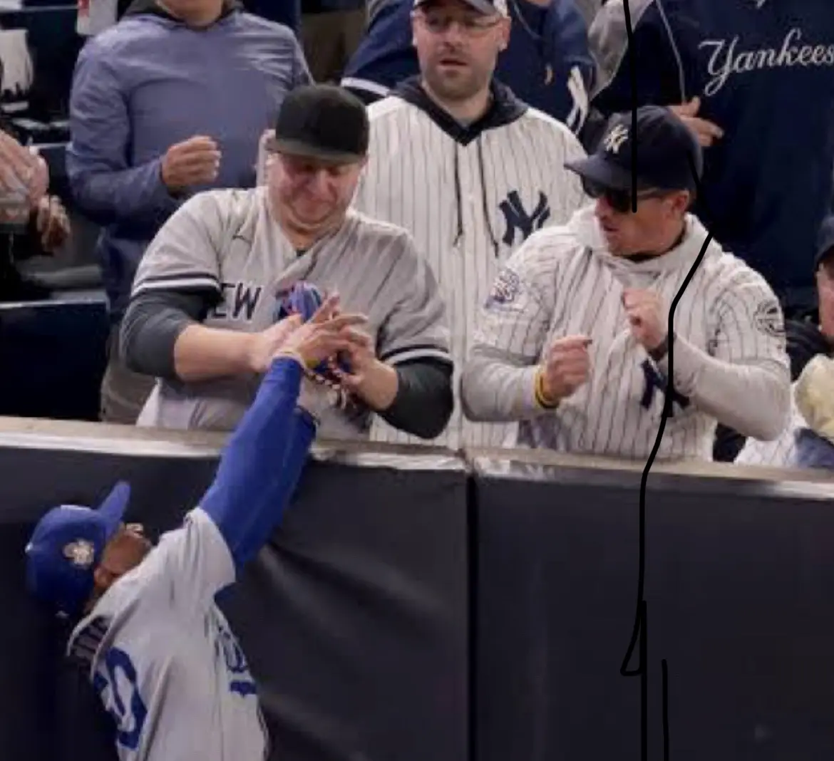 A baseball player in a blue and white uniform reaches over a barrier to high-five a fan in a Yankees jersey. The fan…