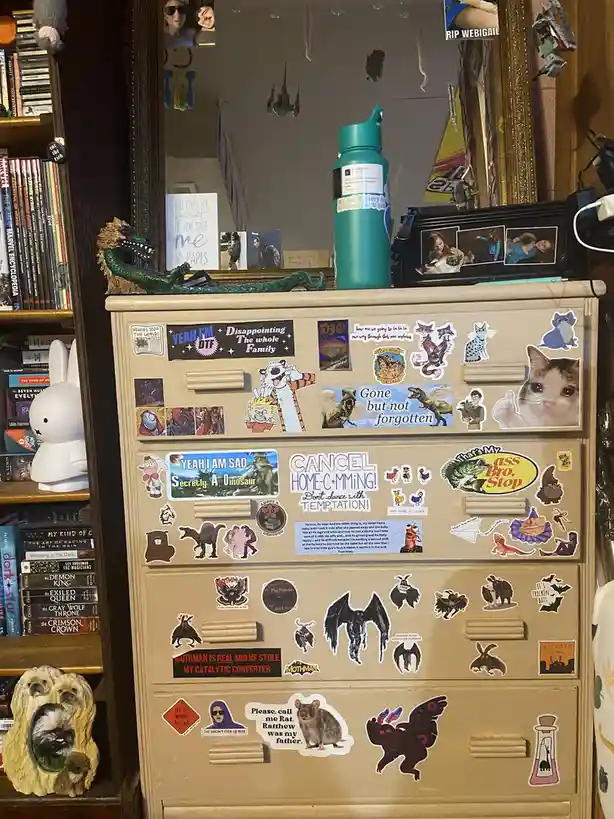 A beige dresser covered in numerous stickers, including cartoon characters, animals, and humorous phrases like 'Cancel…