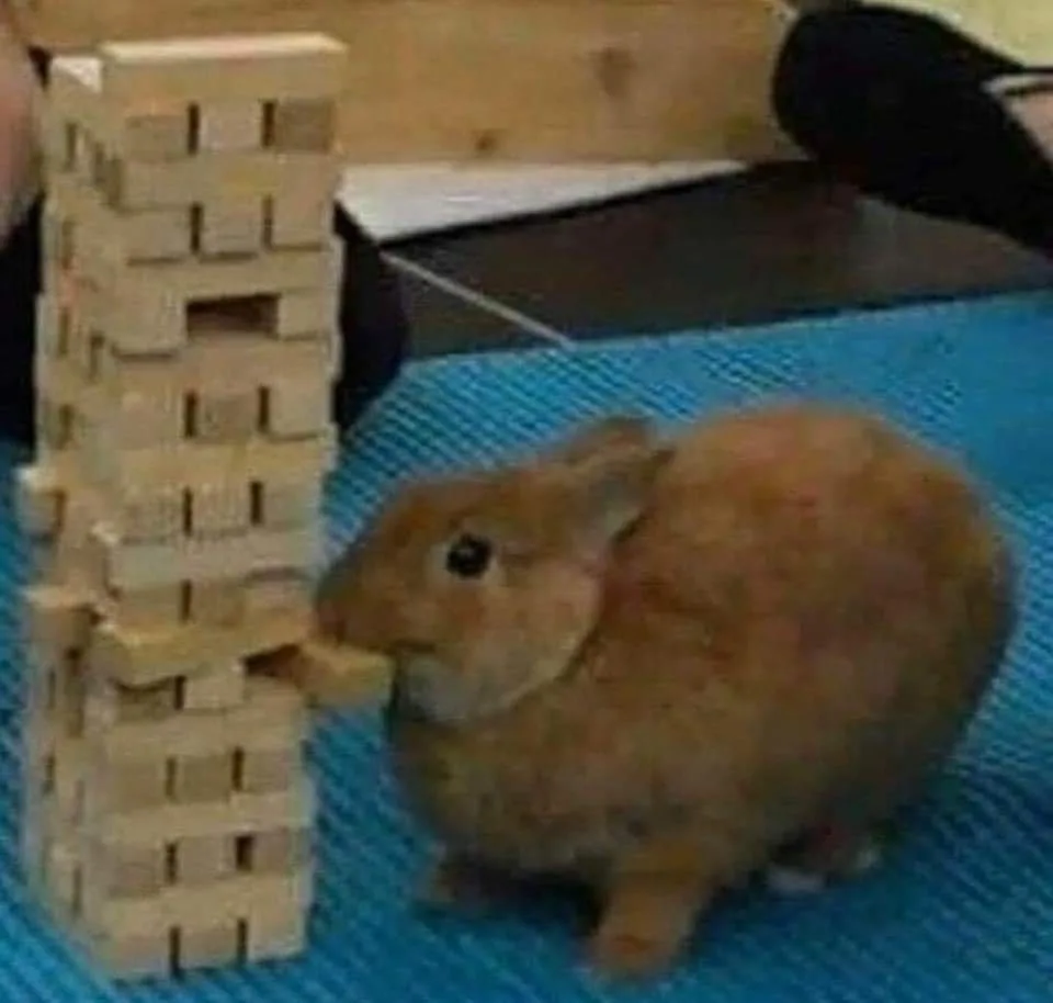 A small brown rabbit sits on a blue textured mat next to a tall, precarious tower of wooden Jenga blocks. The rabbit is…