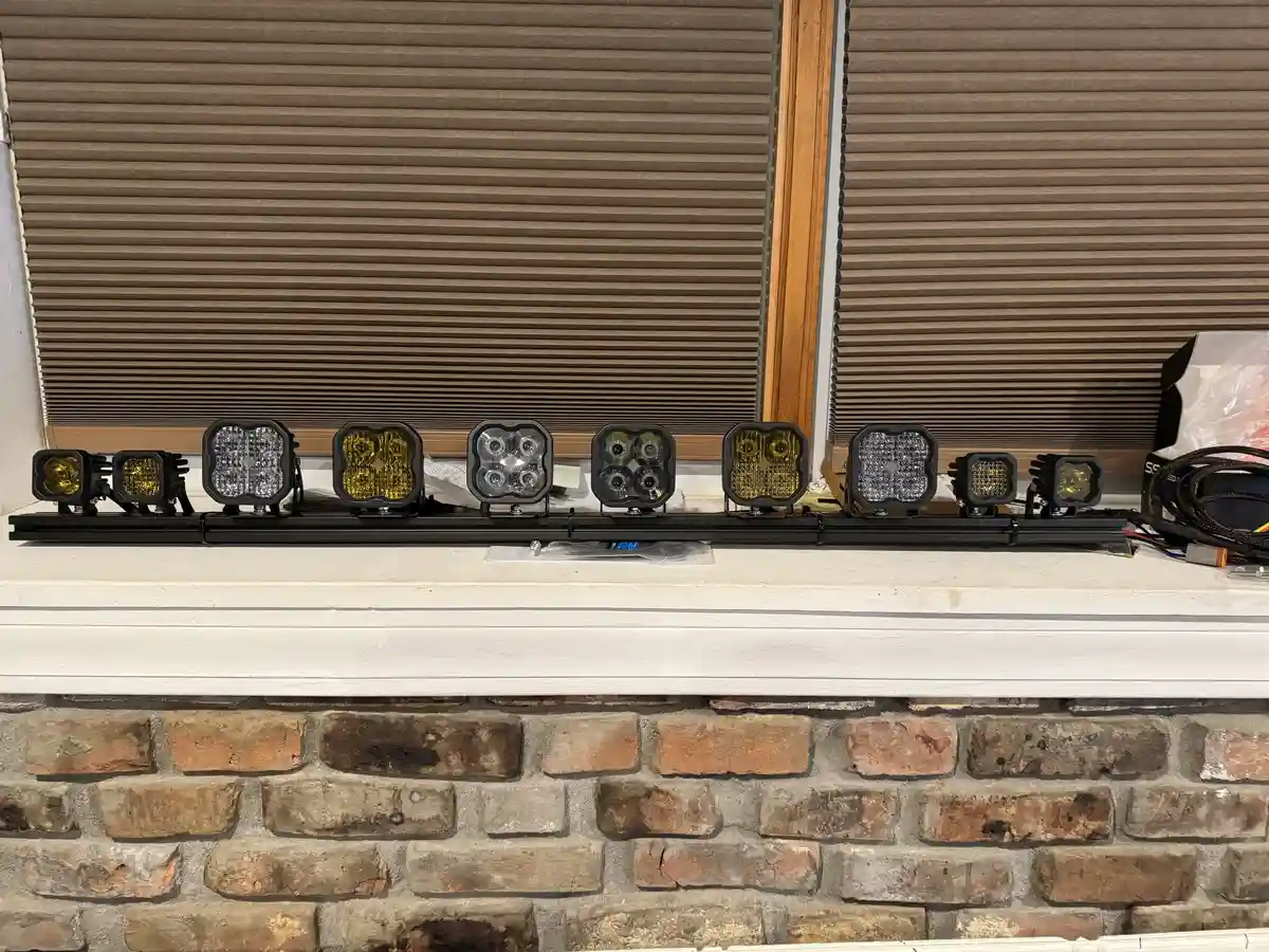 A row of various LED light bars and smaller auxiliary lights are arranged on a white windowsill above a brick wall, with…