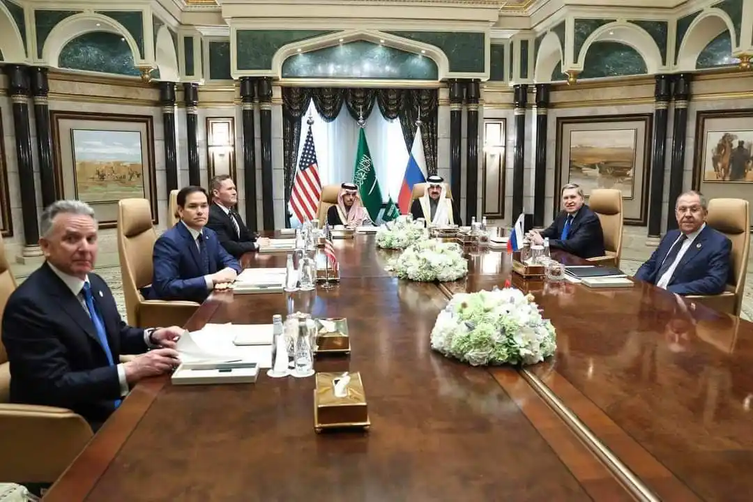a formal diplomatic meeting with several world leaders seated around a long wooden table in an ornate room with flags of the…